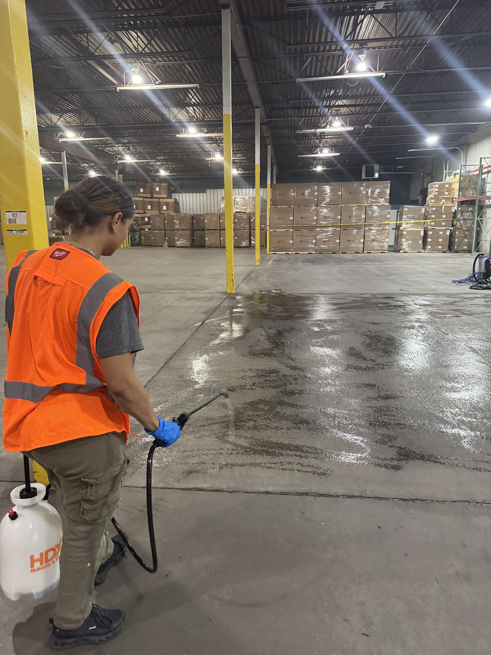 Crew member in a safety vest spraying degreaser on a stained concrete warehouse floor with a pump sprayer.
