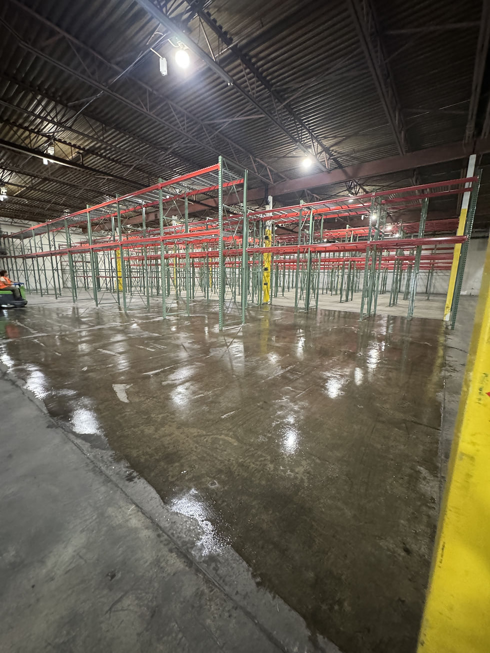 Wet, freshly scrubbed warehouse floor under red and green pallet racking, next to a strip of floor not yet cleaned.