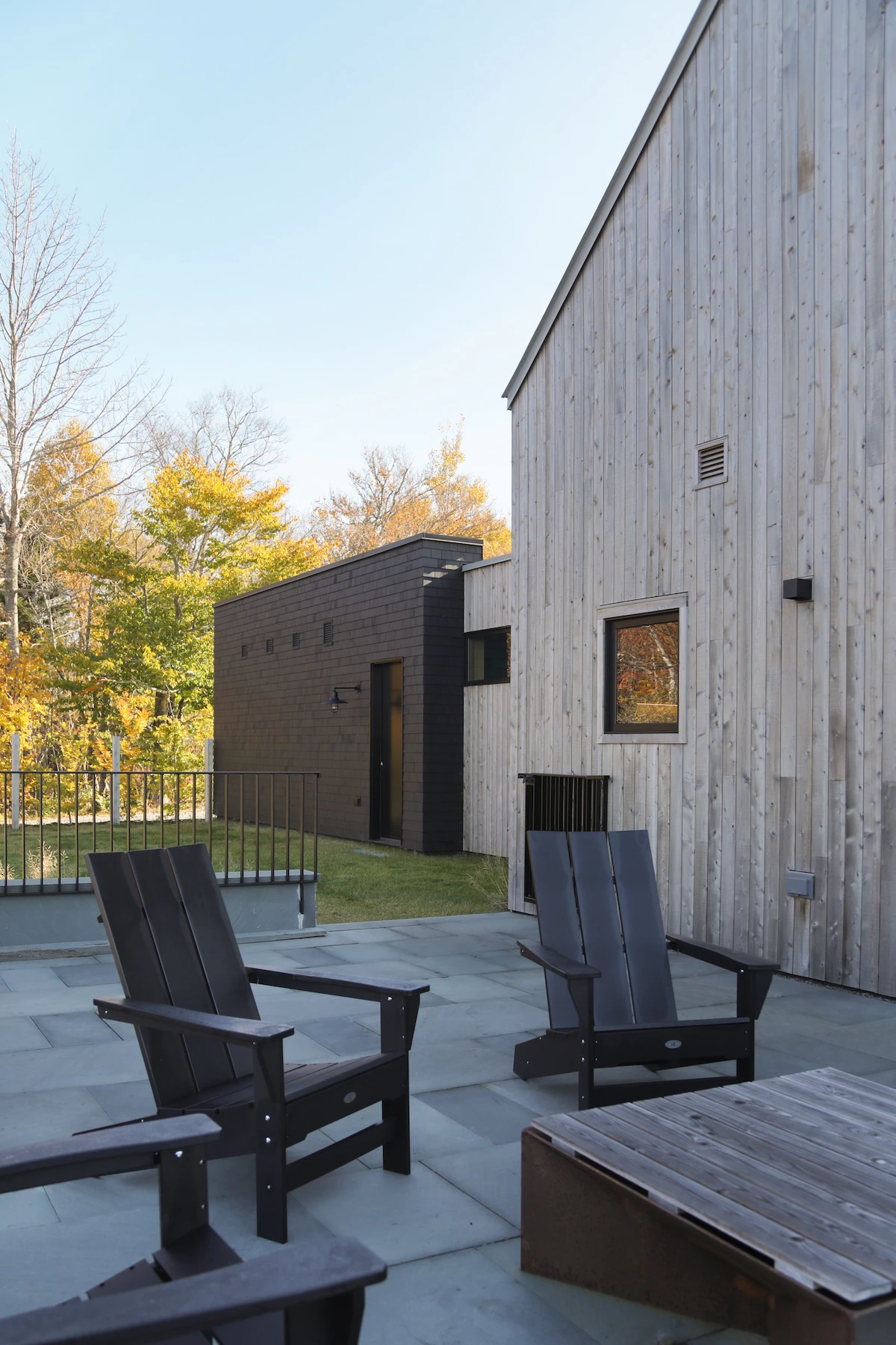 Fire pit on patio with adirondak chairs surrounding. Vertical wood siding on home connected to the garage in the backgroung.