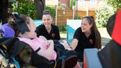 Two caregivers wearing black shirts smile and interact with a person in a wheelchair. The setting is an outdoor patio with sunlit greenery.