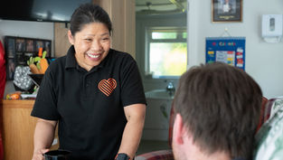 Woman in black shirt with heart logo smiles at seated man, holding mugs. Indoor setting with wall calendar and framed photo in background.