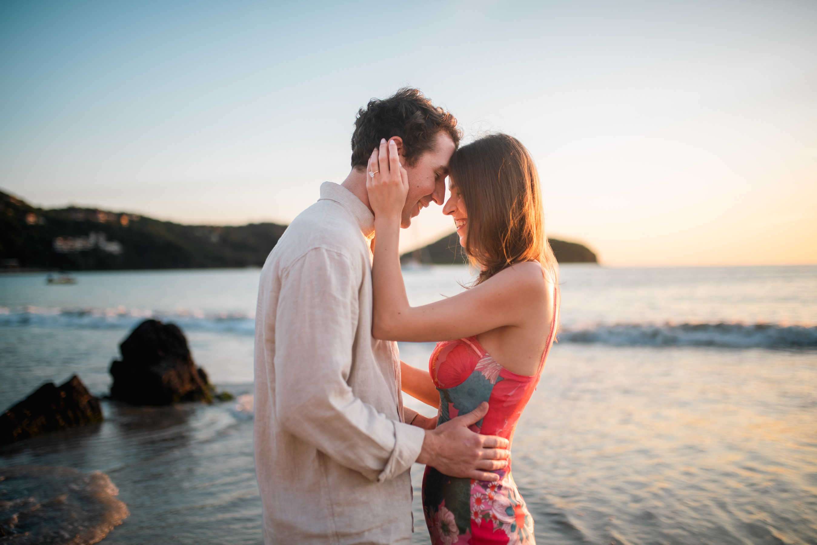 The couple stands on the soft sand of Playa La Ropa, Zihuatanejo, with their foreheads gently touching in a tender moment of intimacy. The golden hues of the sunset surround them, casting a warm glow on their faces. The ocean stretches out behind them, its waves calmly lapping at the shore. Their eyes are closed, savoring the quiet connection, and the scene radiates love, peace, and the promise of a shared future.