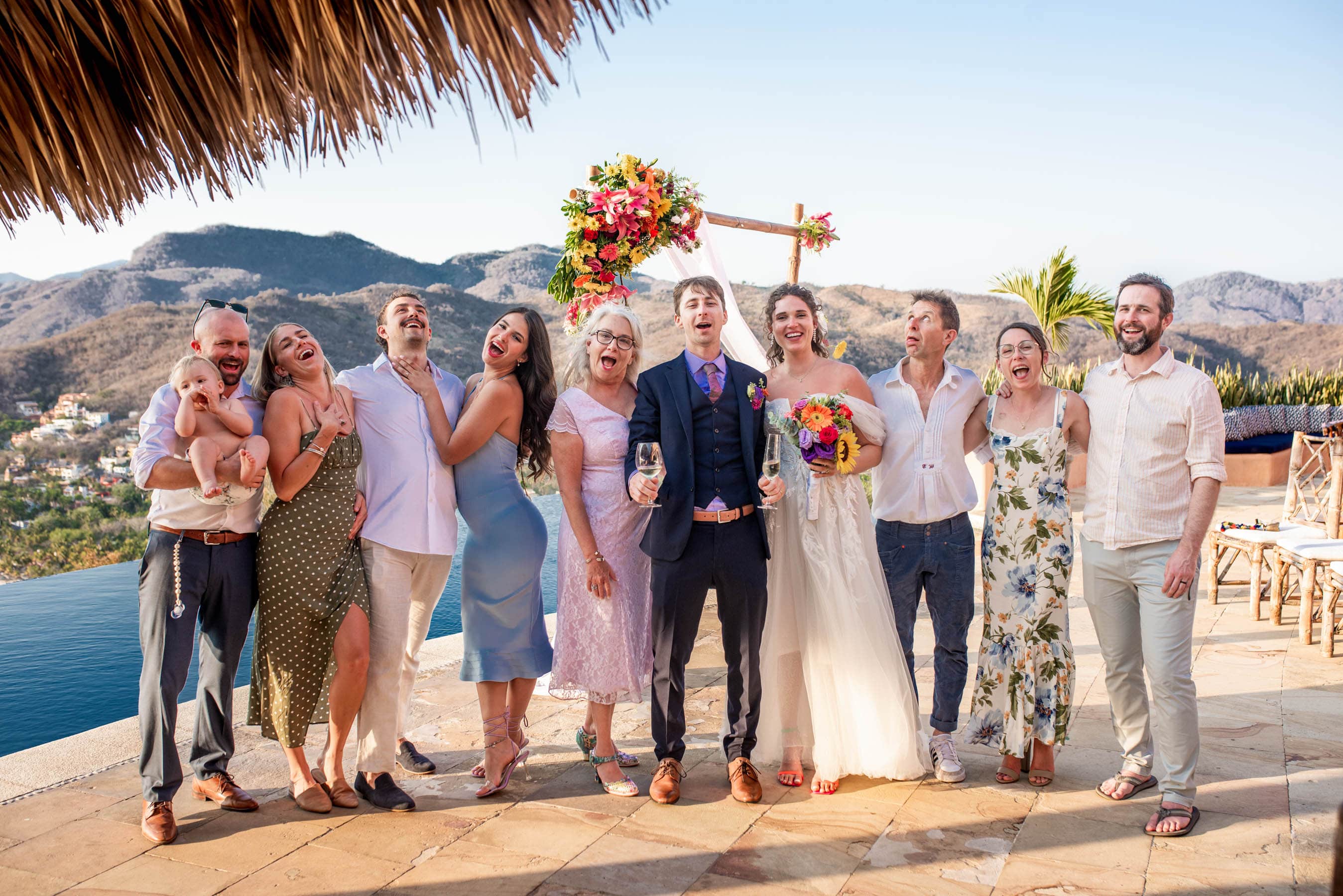 Guests seated on rustic wooden benches facing the ocean, witnessing the heartfelt union of two souls."
"Aerial view of the wedding party gathered on the beach, with colorful parasols providing shade against the bright sun.