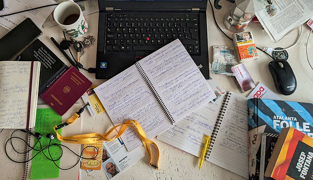 Foto eines Schreibtisches mit Notizheften, Reisepass, Laptop, Büchern, einer Banknote aus Katar