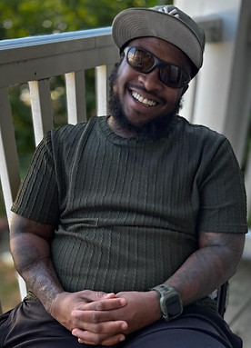 PHOTO OF HUNTA WHO IS A BLACK MAN SITTING OUTSIDE SMILING WITH A WHITE FENCE BEHIND HIM. HE HAVE A BLACK  MUSTACHE AND BEARD, AND HAVE TATTOOS ON BOTH ARMS. HE IS WEARING A FOREST GREEN NEW YORK HAD, BLACK SUNGLASSES, FOREST GREEN LINED T-SHIRT WITH BLACK SHORT, AND FOREST GREEN APPLE WATCH.