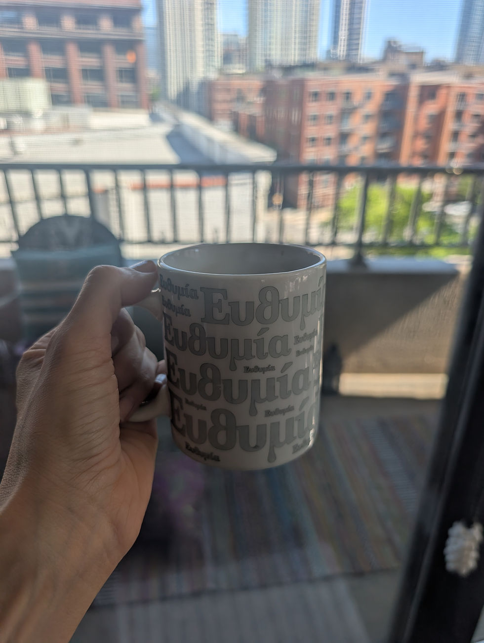 A hand holds a mug with Greek text on it, Ethemia, against a backdrop of a city skyline seen through a balcony.
