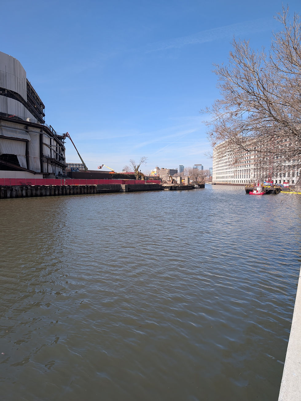Urban river scene with a construction site on the left, a building on the right, leafless trees, and a clear blue sky reflecting on the water.