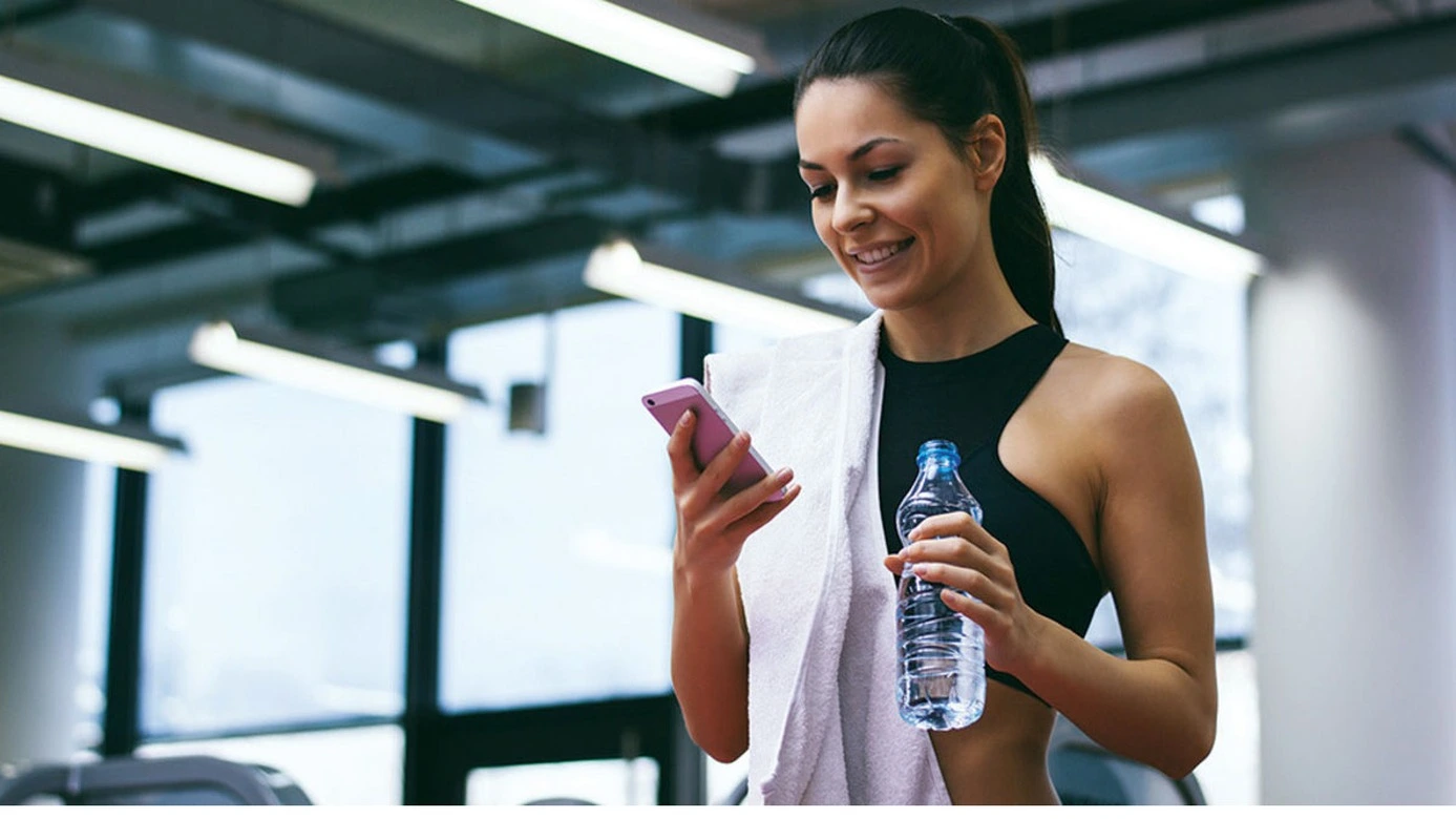 Une femme souriante en train de manipuler son téléphone pour utiliser une application fitness, tout en étant bien préparée pour sa séance. Elle tient une bouteille d'eau dans une main, prête à s'hydrater régulièrement tout au long de son entraînement. Une serviette est soigneusement posée sur son épaule, prête à essuyer la transpiration qui accompagne ses efforts.