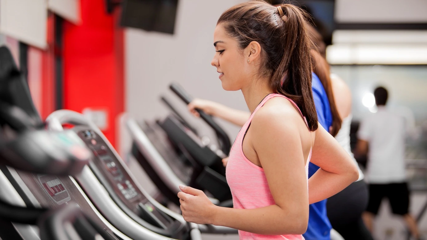 Une jeune femme déterminée, aux cheveux longs, se concentre pleinement et profite intensément de son entraînement sur un tapis roulant dans une salle de sport. Vêtue d'une tenue rouge dynamique, elle incarne la vitalité et la détermination. Ses cheveux flottent gracieusement au rythme de ses pas, ajoutant une touche de fluidité à sa silhouette athlétique.