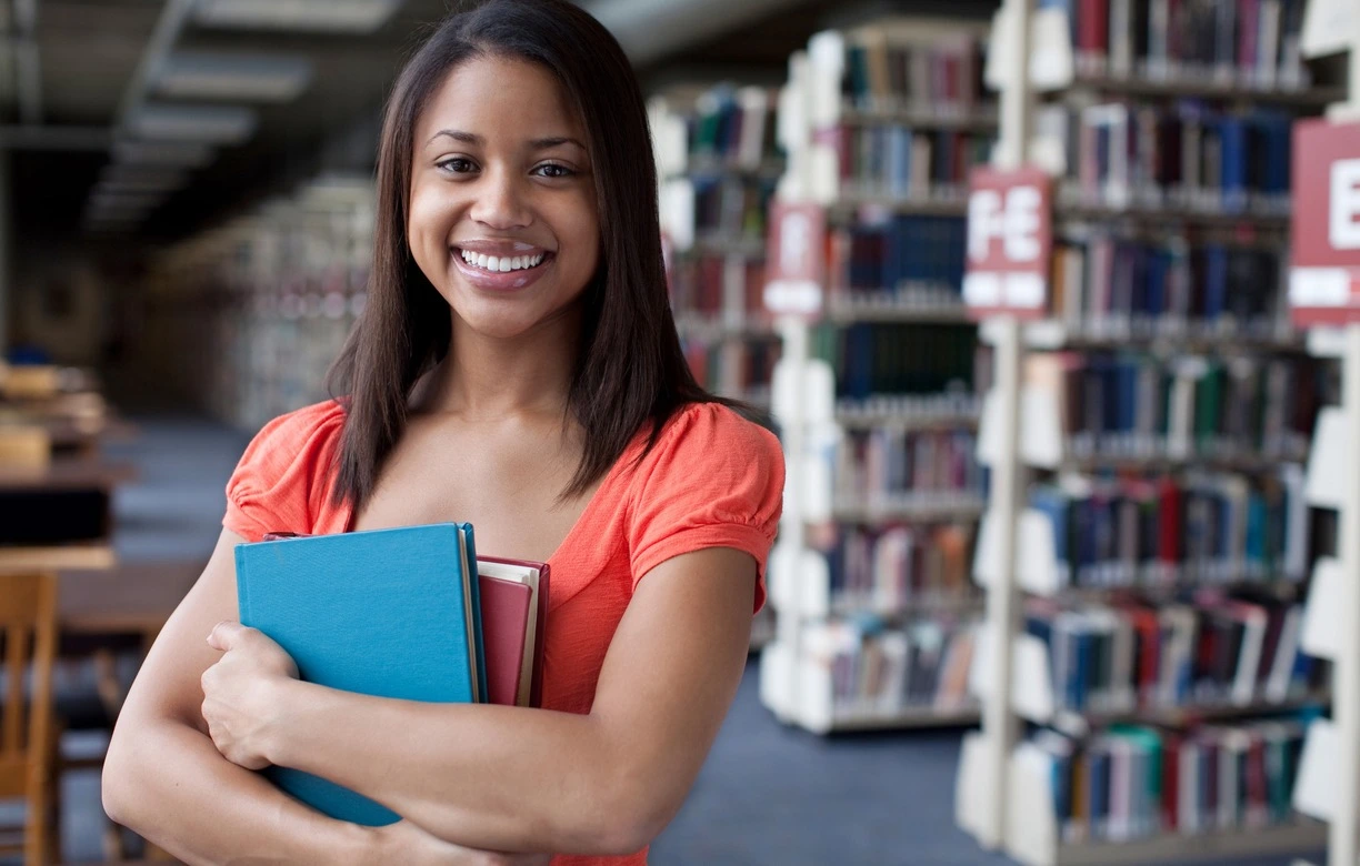 Une étudiante vêtue d'une tenue rouge se trouve dans une bibliothèque de l'université, ses bras encombrés de deux livres qu'elle tient avec enthousiasme. Son visage rayonne d'un sourire radieux, reflétant sa positivité et son bien-être intérieur. Dans cet environnement tranquille et studieux, elle respire la sérénité et l'harmonie.