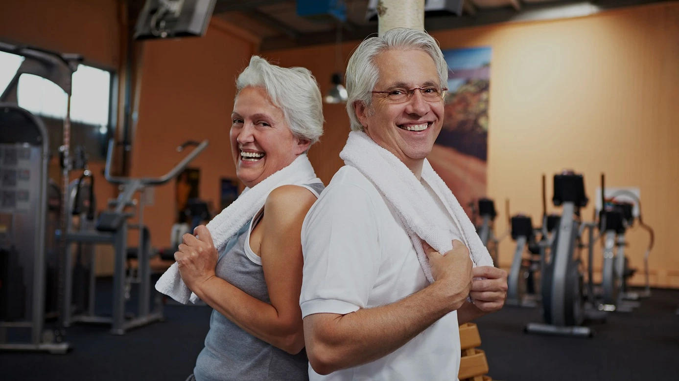 Deux seniors, le visage illuminé par de larges sourires, se tiennent au cœur d'une salle de sport bien équipée, prêts à s'entraîner avec enthousiasme. Ils portent chacun une serviette blanche soigneusement posée sur leurs épaules, symbole de leur engagement envers leur bien-être physique.