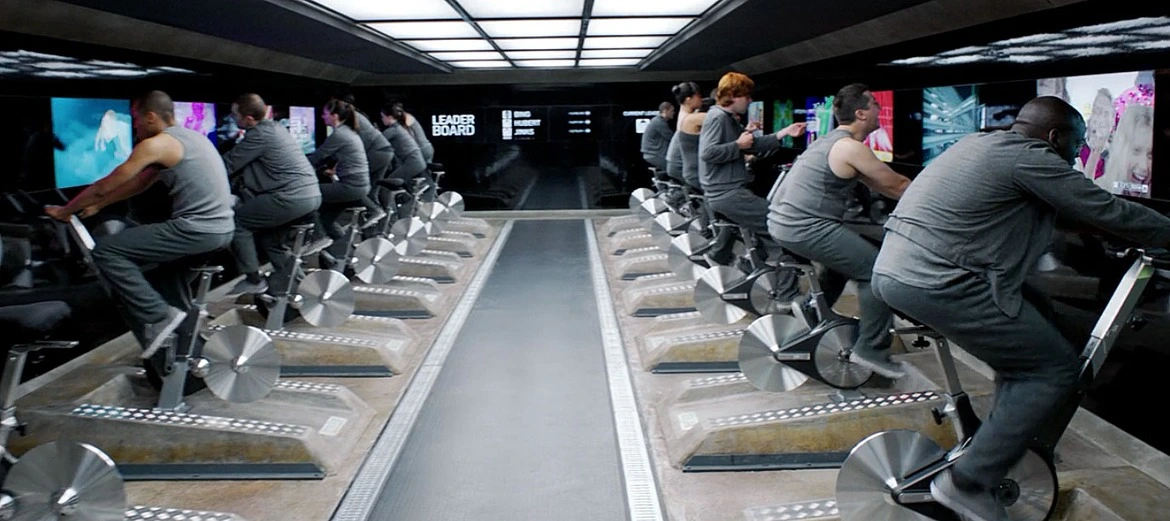 Espace Biking où des personnes vive une expérience immersive. Plus de dix personnes pédalent sur des vélos stationnaires. Les participants sont captivés par ce qui est diffusé sur l' écran devant chacun. Photo de la série Netflix "Black Mirror", Saison 1-Épisode 2.