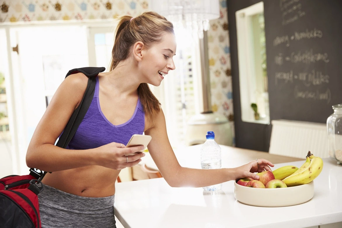 Une femme pleine d'énergie en train de se préparer avant sa séance d'entraînement à la salle de sport. Un sac de sport sur son épaule, un téléphone à la main et l'autre main tient une pomme fraîche, symbole de nutrition saine et de vitalité, démontrant son souci d'adopter de bonnes habitudes alimentaires pour accompagner son activité physique.