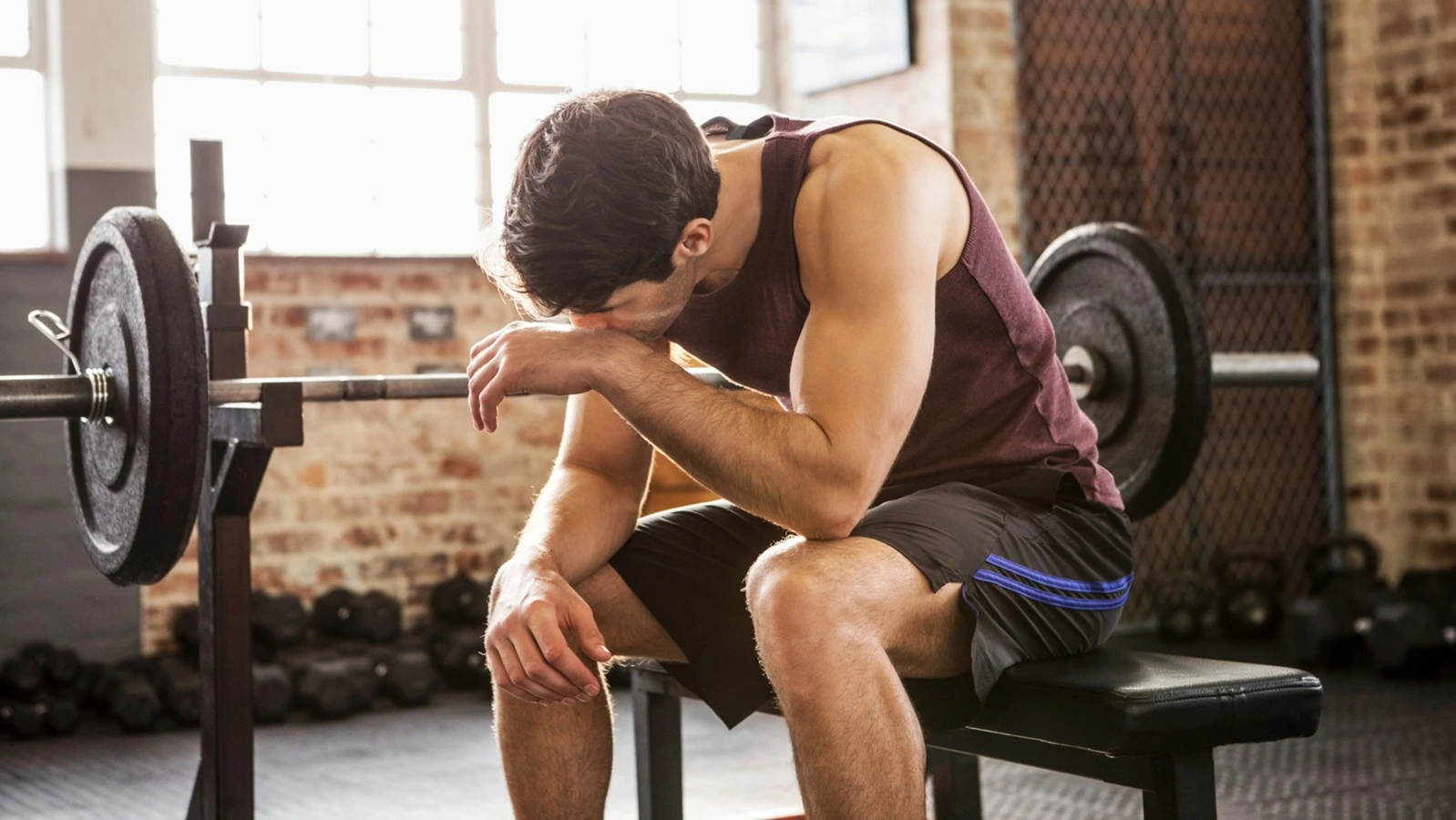 Un homme battu et fatigué s'assoit sur un banc de l'appareil développé couché dans une salle de sport. Son regard perdu dans le vide laisse transparaître une pensée qui le transporte ailleurs. Vêtu d'un t-shirt grenat et d'un short noir. 