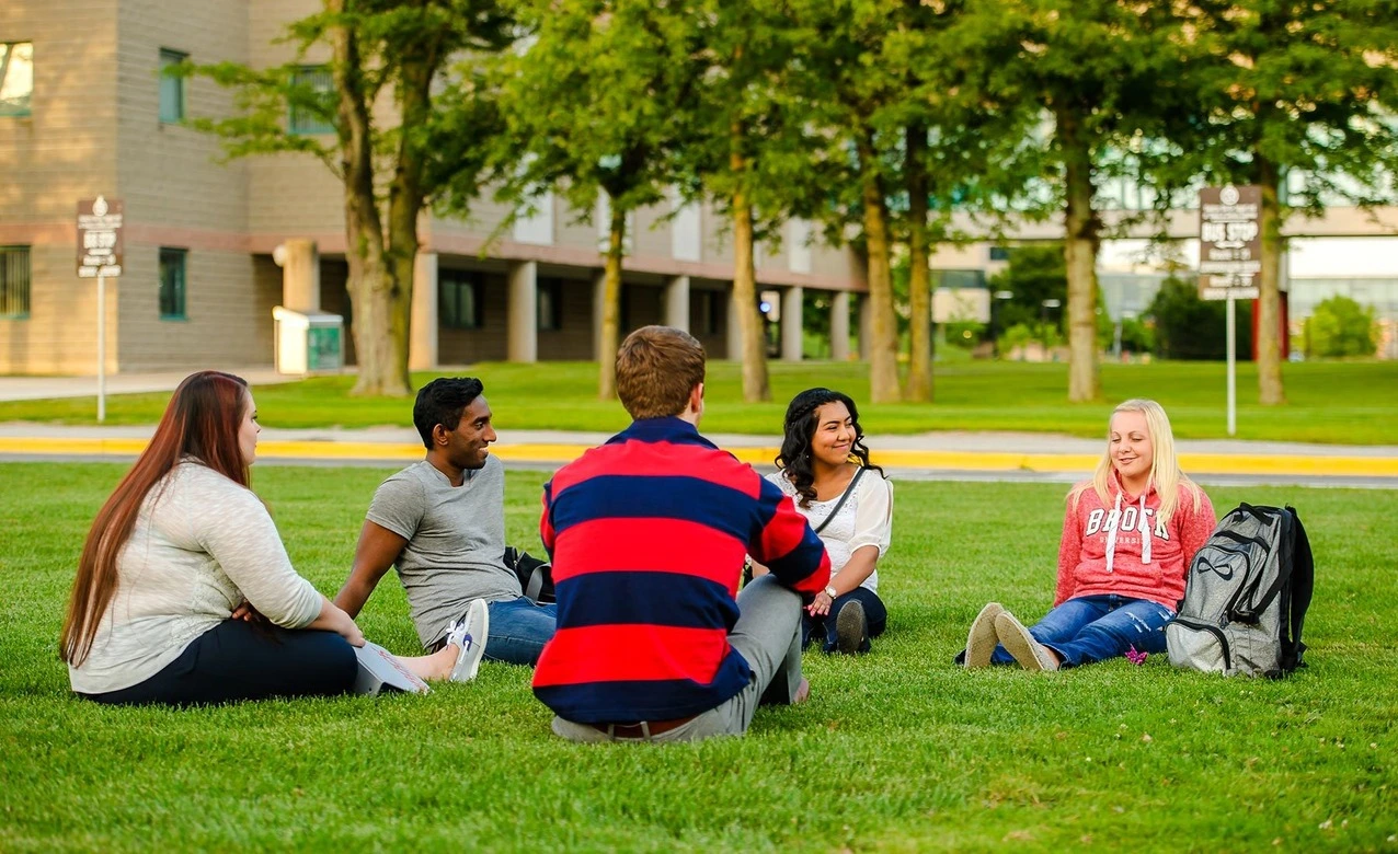 Un groupe chaleureux composé de trois femmes et deux garçons profite du beau temps ensemble dans l'enceinte de l'université. Assis sur l'herbe verdoyante, ils échangent des rires, des discussions animées et des sourires complices. Ces moments de détente renforcent le sentiment d'appartenance à la communauté étudiante et apportent une pause bienvenue dans leur quotidien académique.