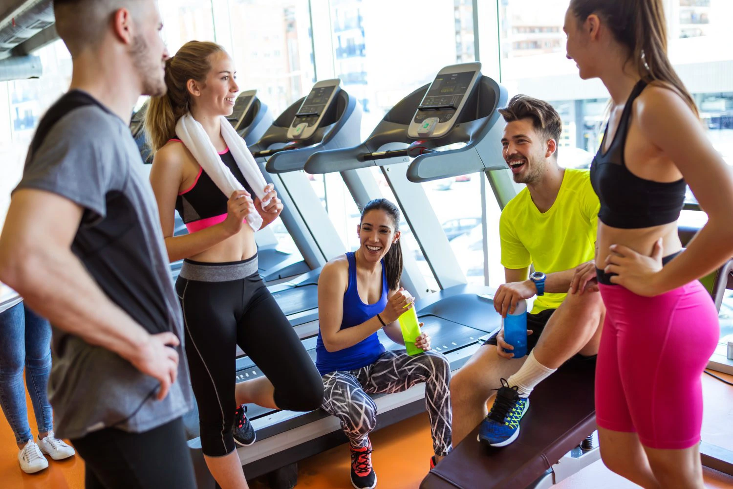 Deux hommes et trois femmes se trouvent dans une salle de sport moderne, baignée de lumière naturelle. Chacun d'entre eux est vêtu de manière appropriée pour une séance de fitness confortable, choisissant des tenues qui leur permettront de rester à l'aise malgré les effets de la canicule. Les hommes portent des hauts de sport techniques et des shorts respirants, tandis que les femmes optent pour des leggings colorés et des débardeurs aérés. Leur choix judicieux de vêtements les aide à rester au frais et à l'aise pendant leur entraînement.