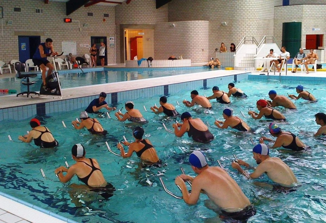 Plus de 10 personnes participent à une séance d'aquabike dirigée par un coach dans une piscine, sous les regards des autres personnes se trouvant au bord de la piscine.