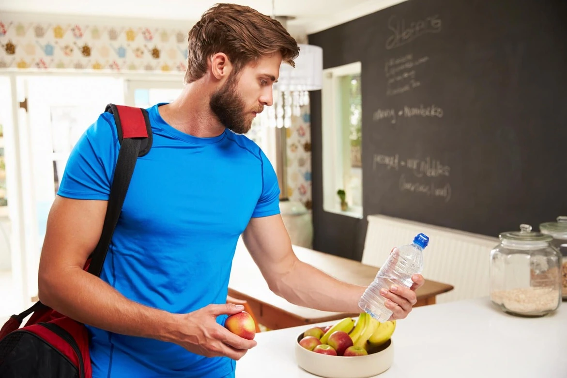 Un homme portant un tee-shirt bleu se prépare à se rendre à la salle de sport. Dans une main, il tient une bouteille d'eau, prêt à rester hydraté pendant son entraînement. Dans l'autre main, il tient une pomme, montrant son engagement envers une alimentation saine. Un sac de sport est négligemment posé sur son épaule, contenant ses affaires pour la séance. Son expression déterminée reflète sa motivation à atteindre ses objectifs de remise en forme.