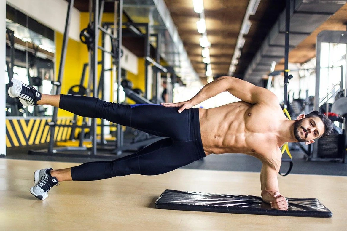 Un homme exécute une planche latérale dans une salle de sport bien équipée. Avec ses abdominaux bien tracés, il maintient une posture stable et solide. Avec son corps aligné en position latérale, il maintient son poids sur l'avant-bras et le côté du pied. Son regard est concentré, démontrant sa détermination à tenir cette position exigeante. Les muscles de son tronc sont sollicités et travaillés intensément dans cet exercice qui vise à renforcer ses abdominaux.