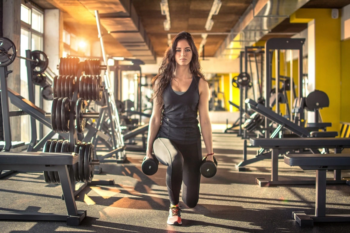 Une femme au regard déterminé et sérieux se trouve dans une salle de sport bien équipée, se concentrant sur le renforcement de sa partie inférieure du corps. Entourée d'équipements spécialisés, elle tient fermement deux haltères dans ses mains. Sa posture est impeccable, ses muscles se contractent avec puissance.