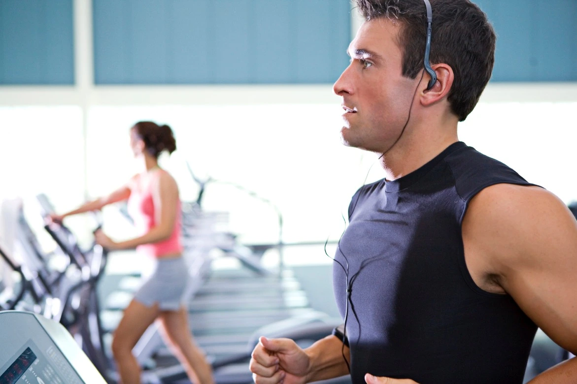 Un homme dynamique s'entraîne avec ardeur sur un tapis roulant dans une salle de sport. Sa silhouette athlétique est en mouvement, ses jambes s'activant. Un léger sourire se dessine sur son visage alors qu'il court, porté par la musique qui résonne dans ses écouteurs.