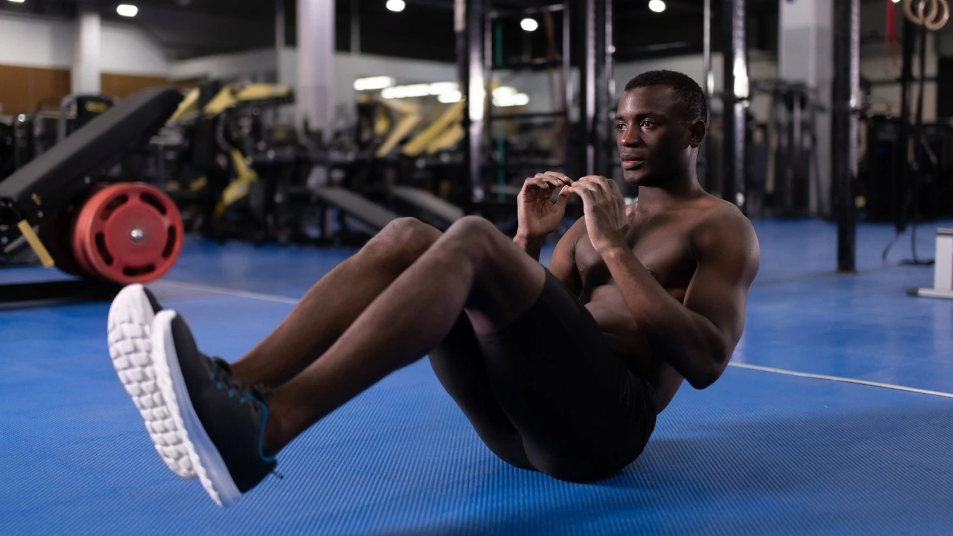 Un homme déterminé et concentré s'entraîne seul dans une salle de sport bien équipée. Vêtu d'un short noir et d'un débardeur, il se tient au centre de la salle, entouré d'appareils de musculation et de tapis d'exercice. Son regard est fixé sur l'objectif de renforcer son core. Chaque mouvement est exécuté avec précision, mettant en évidence sa forme physique et sa détermination.