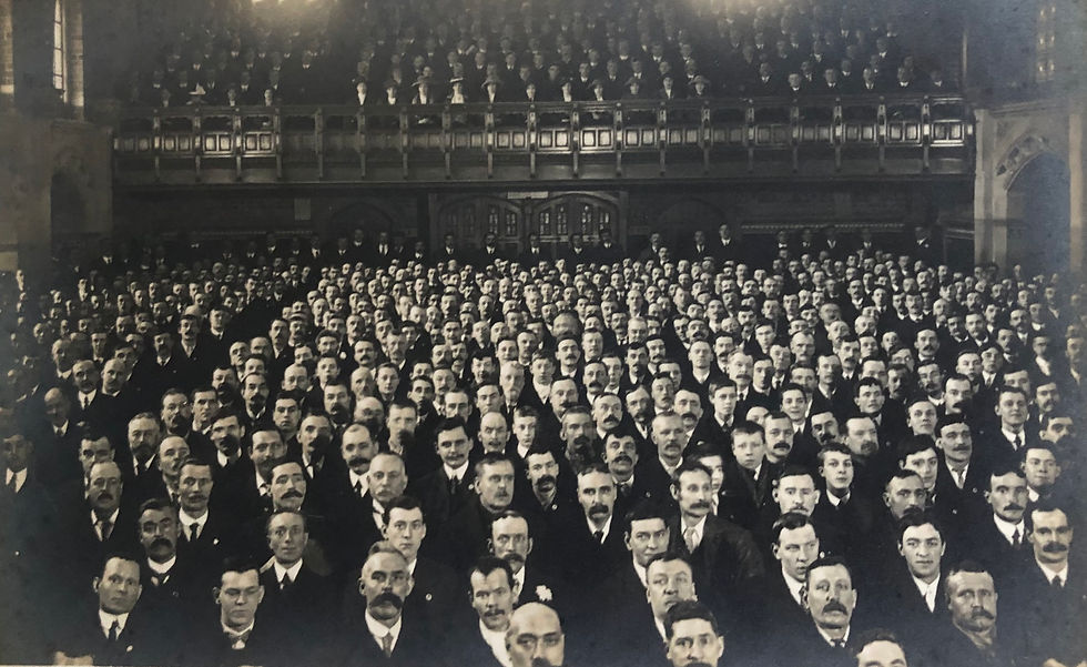 A black and white vintage image shows row after row of men sitting in a gothic-style town hall. They are looking at the camera. behind them can be seen some women and more men in seats in a balcony.