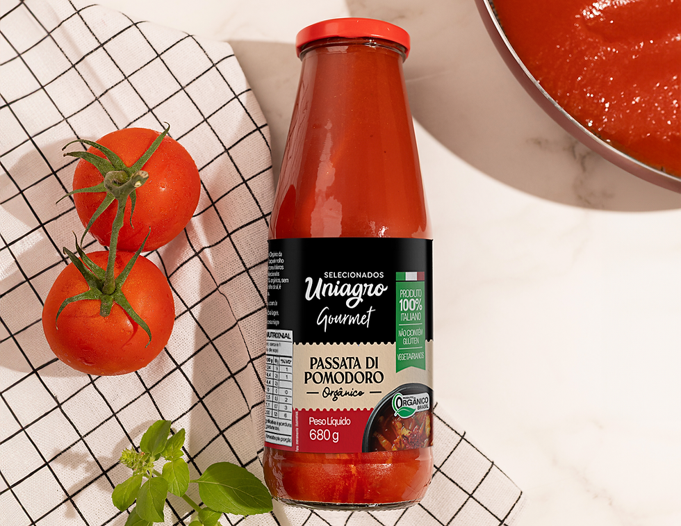 Garrafa de passata de tomate orgânica com rótulo preto, ao lado de tomates frescos sobre pano xadrez; molho em panela ao fundo.