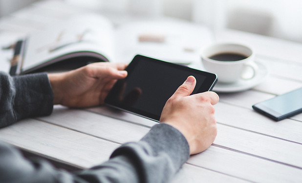 Hands holding a small tablet horizontally at a table with a book, coffee, and phone