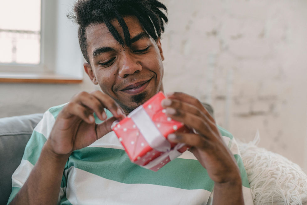 Man in a striped shirt happily examines a small red polka dot gift box. He is seated on a couch in a softly lit room with a light background.
