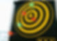 Yellow and black dartboard with red and green darts stuck in various rings, displaying scores from 10 to 100. Bright, blurred background.