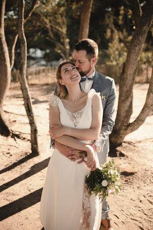 Dans un petit bois, au bord de la mer, le marie se tient debout derriere la mariee, la serre contre lui et l'embrasse sur la joue 