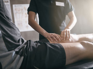 Therapist in black shirt massages a patient’s leg on a treatment table in a bright clinic.