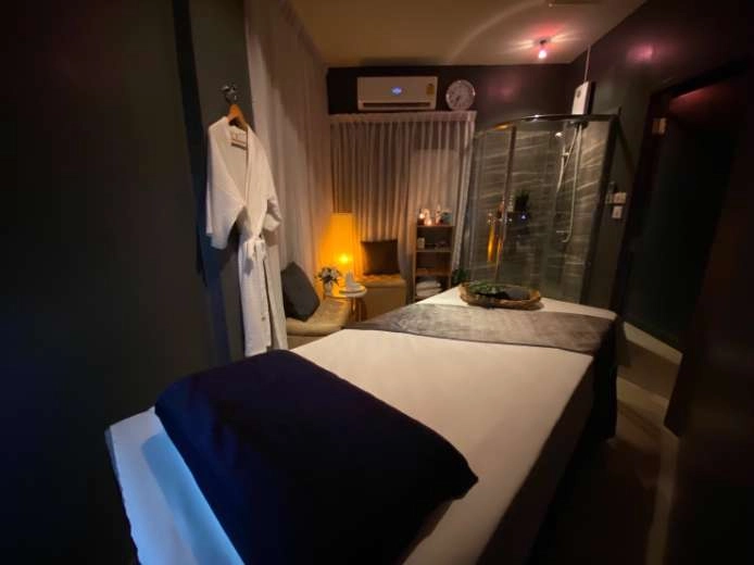 A dimly lit spa room at S'Sense Massage, one of the most trusted gay massage Bangkok destinations, featuring a massage table covered in white and gray linens. A robe hangs on the wall, and a cozy seating area with soft lighting creates a serene and welcoming atmosphere.