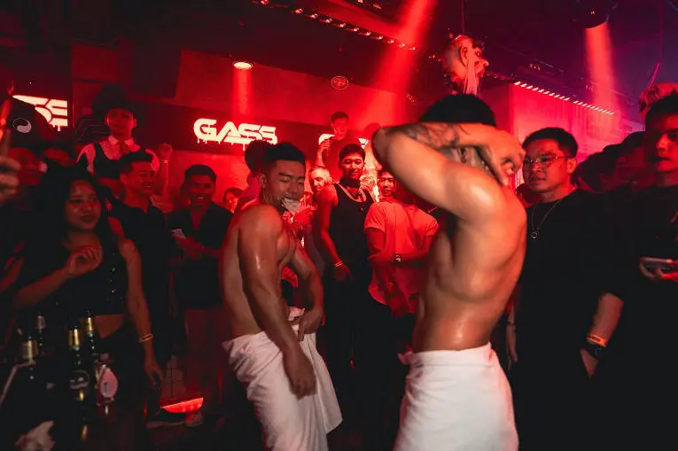 In the electrifying heart of gay Bangkok during a iconic towel-themed night at a massive circuit party, a shirtless man wrapped in a crisp white towel dances with unbridled joy alongside a group of equally enthusiastic men, fueling the high-energy and carefree vibe of the lively celebration.