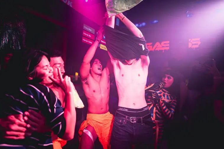 At a lively outdoor gathering in gay Bangkok, a shirtless man proudly holds up a bag of vibrant green goodies, flashing a confident smile directly at the camera amid the relaxed and festive casual vibe.