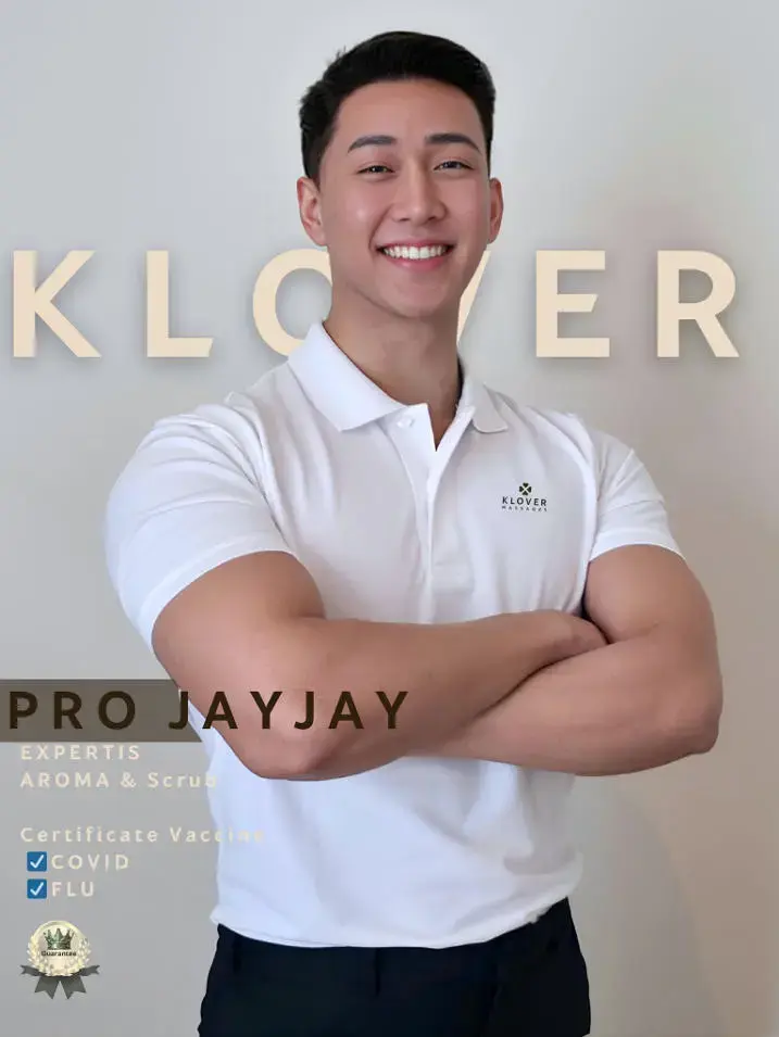 In the welcoming world of gay Bangkok's premium men's massage spas, professional therapist Pro JayJay beams with a confident smile in a crisp white polo shirt featuring the "Klover" logo, standing with arms crossed against a simple beige wall, highlighted by text promoting his expertise in Aroma & Scrub treatments and certified vaccinations for COVID and FLU.