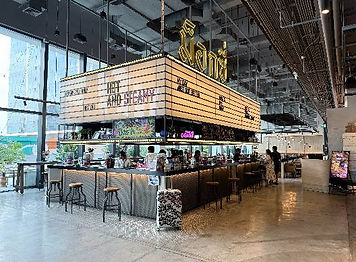 Modern indoor bar at a gay hotel in Bangkok, featuring high stools, a large illuminated marquee overhead, and floor-to-ceiling windows. A few people are seated at the bar and walking nearby.