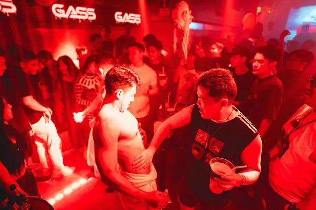 At a thrilling gay Bangkok nightlife event, a charismatic shirtless man takes center stage, passionately addressing the crowd while standing prominently in front of an attentive and energized audience.