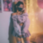 Woman in off-shoulder top, pleated skirt, and mask leans against a wall with art.