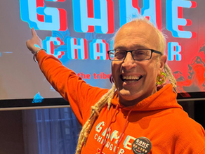 Alex Roland smiling and wearing a orange hoodie stood in front of a screen showing the Game Changer logo.