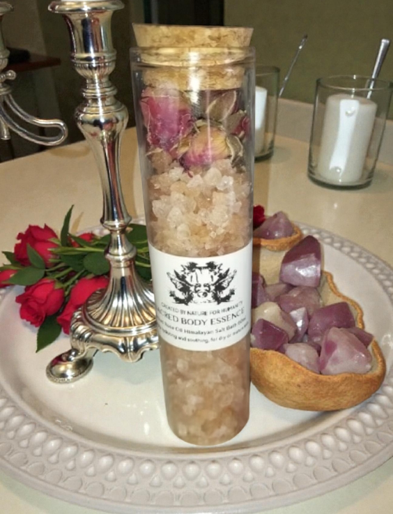 Glass bottle labeled "Sacred Body Essence" with Himalayan salt and dried roses, set on a tray with red roses, candle holders, and crystals.