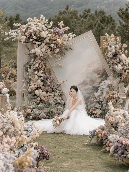 Bride seated among soft Morandi wedding florals with a painted backdrop, pastel garden installation, and romantic fine art bridal styling.