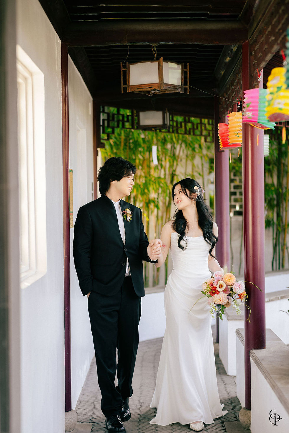 Bride and groom holding hands and smiling at each other beneath colorful Chinese lanterns at Dr. Sun Yat-Sen Classical Chinese Garden in Vancouver, surrounded by traditional red wooden pillars, bamboo, and elegant Chinese garden architecture.