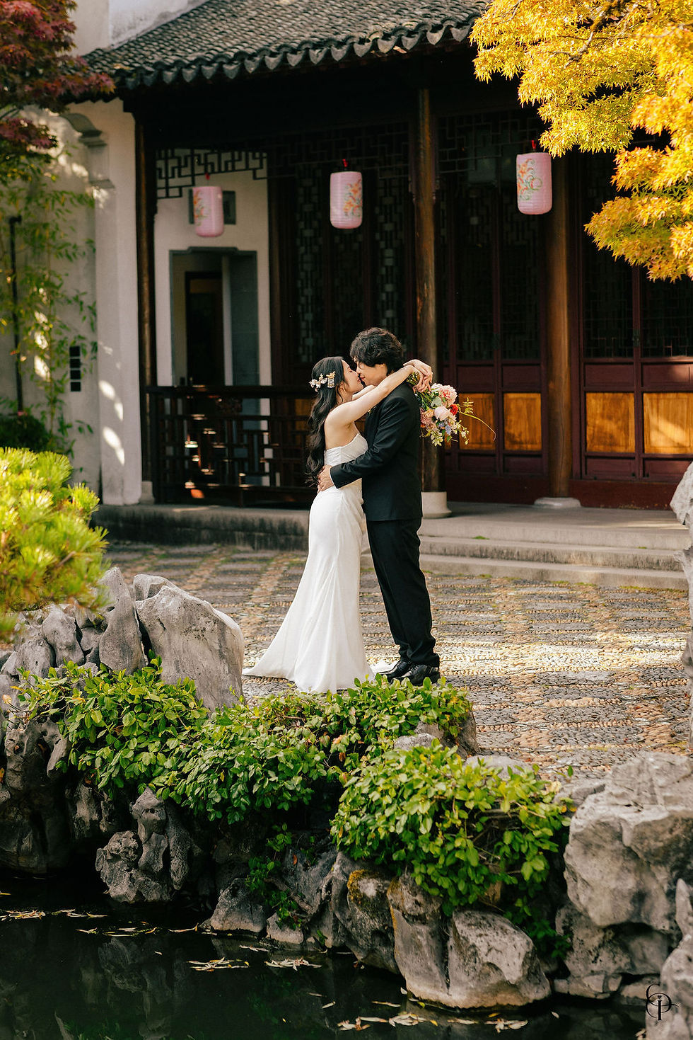 Bride and groom sharing a romantic kiss in the courtyard of Dr. Sun Yat-Sen Classical Chinese Garden in Vancouver, surrounded by traditional Chinese wooden architecture, natural stone pathways, lush greenery, and warm afternoon sunlight.