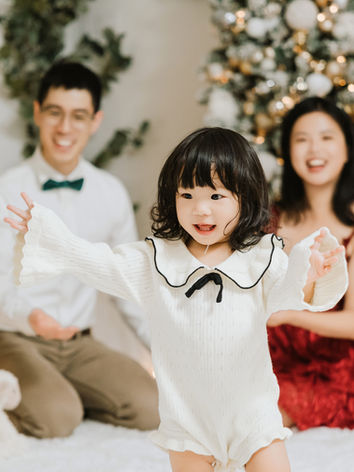 Little girl in a white knit outfit playing in front of her smiling parents during a Vancouver Christmas family photoshoot, with a glowing fireplace and decorated Christmas trees in the background.