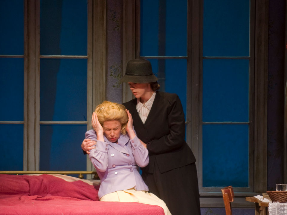 Photograph of a middle aged blonde woman in 1940's attire and hairstyling, covering her ears and sitting on a bed while a younger woman in a severe blue suit and hat grabs her shoulders.