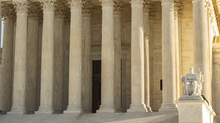 Exterior of a grand neoclassical building, featuring multiple tall white Corinthian columns with ornate capitals. Marble steps lead up to the columns, with a stone sculpture visible to the right side. The columns have deep vertical grooves and stand in front of a light-colored stone facade, representing a judicial or governmental building.