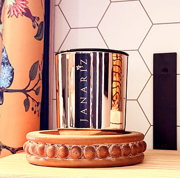 Rose gold jar Janariz Cashmere Vanilla scented  luxury candle with vertical black and gold label, displayed on a round terracotta tray in front of a white hexagon tile wall and floral curtain.