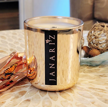 Rose gold jar Janariz Aqua de lluvia scented luxury candle with vertical black and rose gold label, displayed on a marble table next to a rose gold lid with a butterfly design and decorative wicker balls.